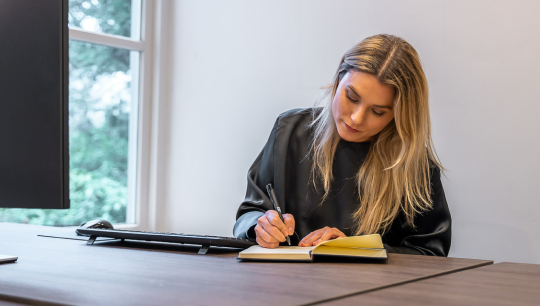 Vrouw schrijft in haar boekje achter een bureau
